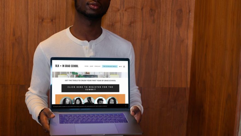 A black man wearing a long sleeved white Henley shirt holds an open laptop  against his chest presenting the screen toward the camera. On the laptop screen is information about the Grad <a href=
