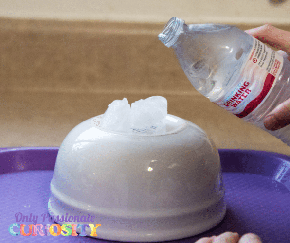 White ceramic bowl turned upside-down with ice cubes on top. A person is pouring water onto the ice from a plastic bottle.